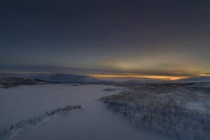 Et rolig, snødekt landskap i skumringen med mørke, bølgende åser i bakgrunnen. Himmelen går over fra dyp blå til en svak gul i horisonten, og kaster en myk, gylden glød over den vinterlige scenen. Sparsom vegetasjon titter gjennom snøen.