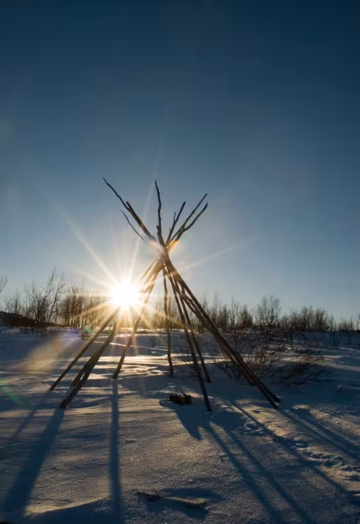 En tradisjonell samisk lavvu står i et snølandskap med sparsomme trær i bakgrunnen. Solen skinner sterkt gjennom trebjelkene, og kaster lange skygger på den snødekte bakken under en klarblå himmel.