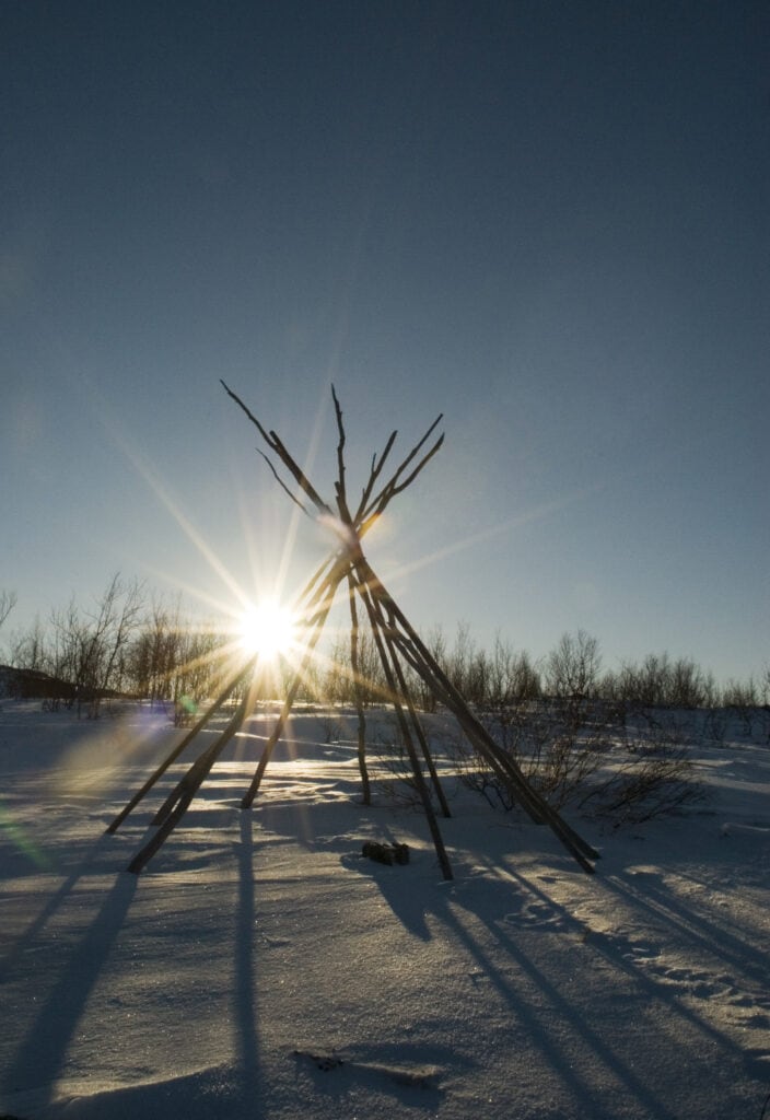En tradisjonell samisk lavvu står i et snølandskap med sparsomme trær i bakgrunnen. Solen skinner sterkt gjennom trebjelkene, og kaster lange skygger på den snødekte bakken under en klarblå himmel.