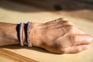 Et nærbilde av en hånd som hviler på en treoverflate, iført to vevde armbånd. Armbåndene har intrikate mønstre, som hver består av metalliske tråder sammenflettet med tråder i forskjellige farger. Huden er opplyst av naturlig sollys, og kaster myke skygger.