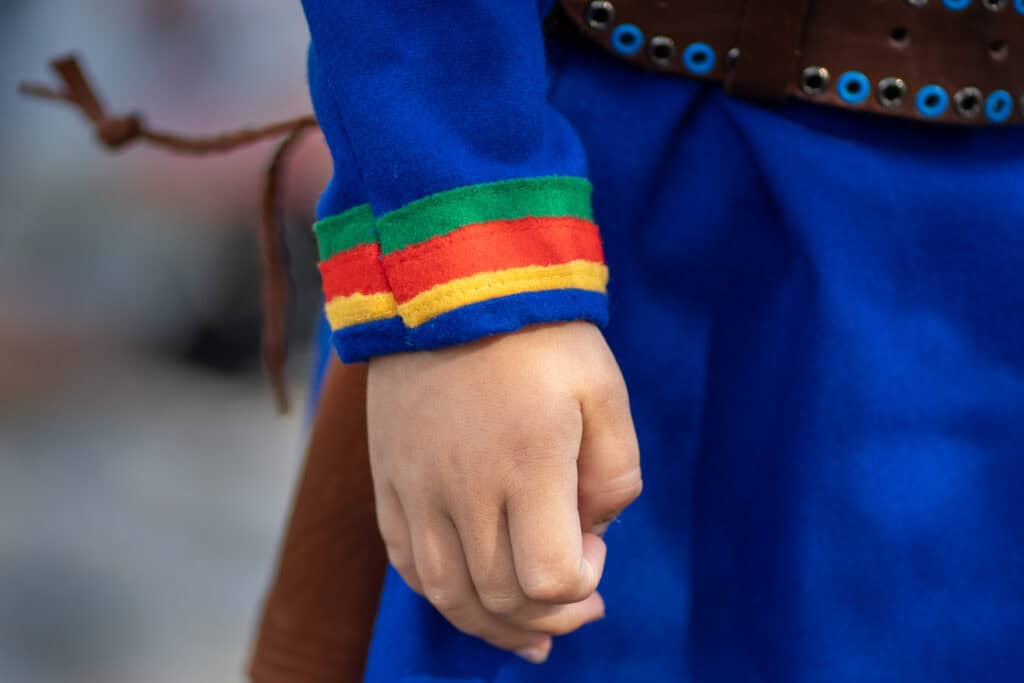 Nærbilde av en barnehånd iført et blått langermet plagg utsmykket med fargerike striper av grønt, rødt og gult nær mansjetten. Antrekket inkluderer også et belte med sirkulære dekorasjoner. Bakgrunnen er uskarp.