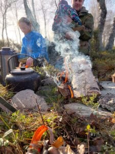 En person i en blå jakke pleier en vannkoker på et bål, mens en annen person i en kamuflasjejakke holder en baby. Røyk stiger opp i skogmiljø med tømmerstokker og spredt løvverk rundt.