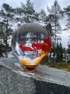 En kule av glass med røde reinsdyrdesigner hviler på en stein, satt mot et skogbakgrunn med høye trær og overskyet himmel. Kulen reflekterer de naturlige omgivelsene, og skaper en harmonisk blanding av kunst og natur.