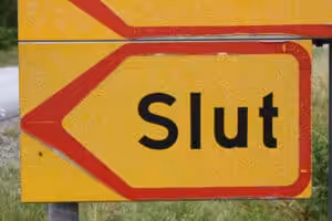 Et gult veiskilt med røde kanter viser ordet "Slut" med fete, svarte bokstaver. Bakgrunnen viser et gresskledd område, noe som antyder landlige omgivelser eller veikanter.