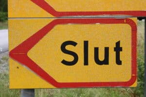 Et gult veiskilt med røde kanter viser ordet "Slut" med fete, svarte bokstaver. Bakgrunnen viser et gresskledd område, noe som antyder landlige omgivelser eller veikanter.